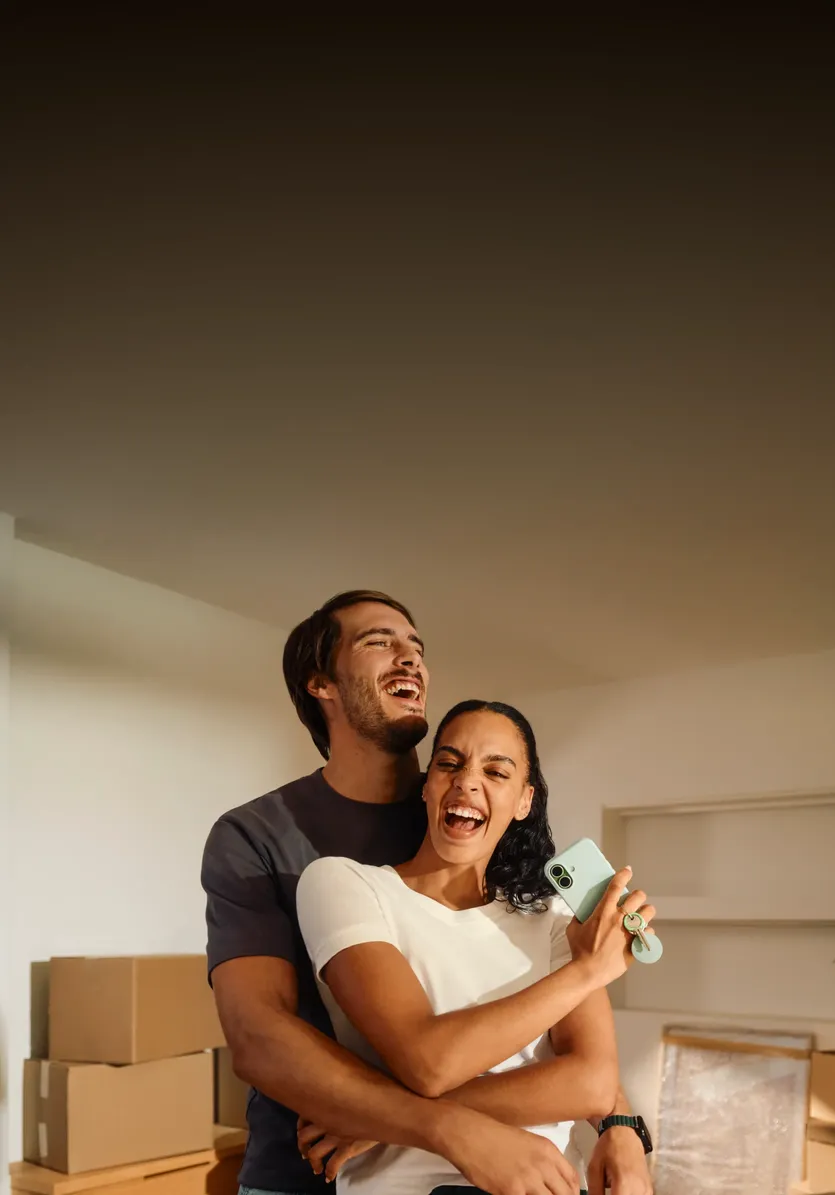 A smiling man embraces a woman from behind as she laughs and holds a smartphone with house keys inside an apartment with cardboard moving boxes.