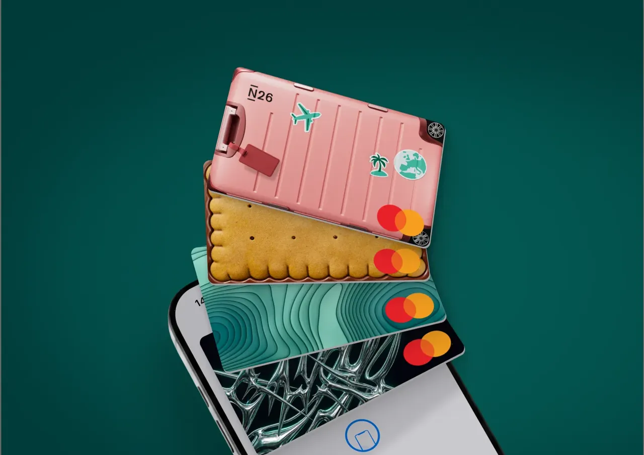 Vier individuell gestaltete N26-Kartendesigns, darunter eines im Stil eines rosa Koffers und eines wie ein Keks, fächern sich vertikal über einem Smartphone-Bildschirm auf.