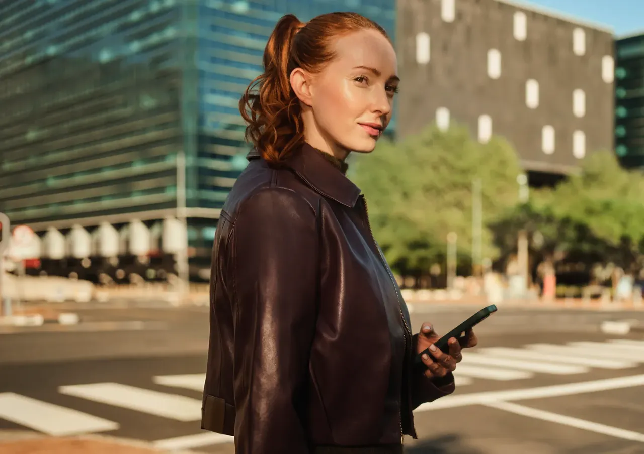 Eine Frau mit einem zu einem Pferdeschwanz gebundenen roten Haar und einer braunen Lederjacke steht an einem städtischen Fußgängerüberweg und hält ein Smartphone in der Hand.