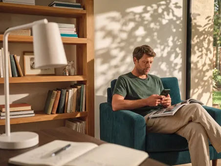 A man sits in an armchair next to a bookshelf, using his smartphone with an open magazine on his lap.
