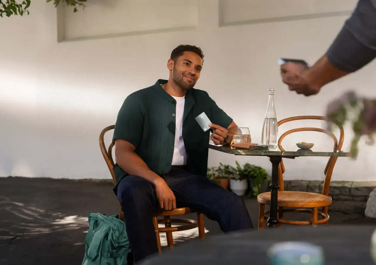 Ein an einem Tisch im Außenbereich eines Cafés sitzender Mann lächelt, während er eine graue Zahlungskarte hochhält.