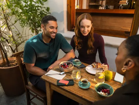 A group of friends sits around an outdoor cafe table enjoying drinks and breakfast bowls with a payment card resting on a phone nearby.
