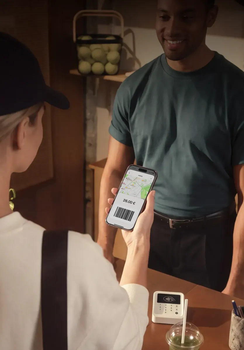 Eine Person von hinten hält einem Kassierer ein Smartphone mit einem Barcode und einer Kartenansicht entgegen, während zentral das weiße N26-Logo eingeblendet ist.