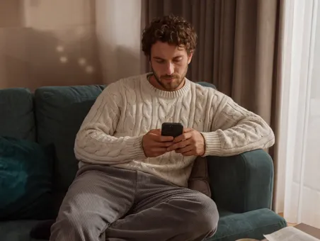 A man wearing a knit sweater sits on a blue couch using his smartphone.