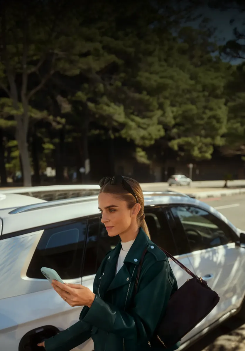Une femme en veste vert foncé tenant un smartphone se tient à côté d'une voiture blanche garée près d'une rue bordée d'arbres.