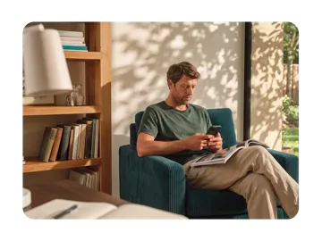 A man sits in a teal armchair using his smartphone while holding an open magazine in a brightly lit living room next to a wooden bookshelf.