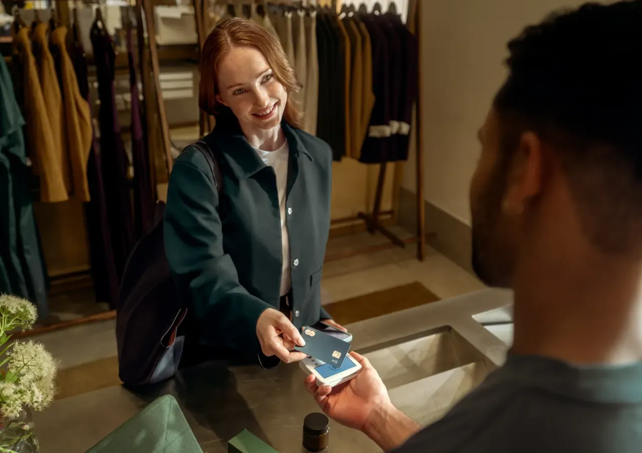 Eine blinde Frau mit Pferdeschwanz von hinten zeigt einem Mitarbeiter an der Kasse einen Smartphone-Bildschirm mit einem Barcode und dem Betrag von 26,00 €.