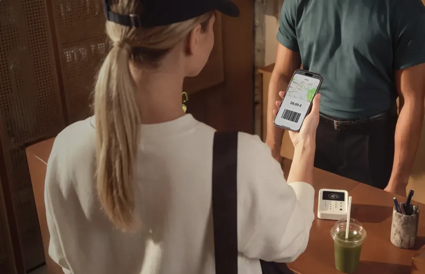 A woman with blonde hair in a ponytail seen from behind shows a smartphone screen displaying a barcode and a total of 26.00 € to a store employee at a checkout counter.