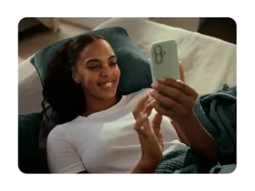A smiling woman lies on a bed with her head resting on a pillow while looking at her smartphone screen.