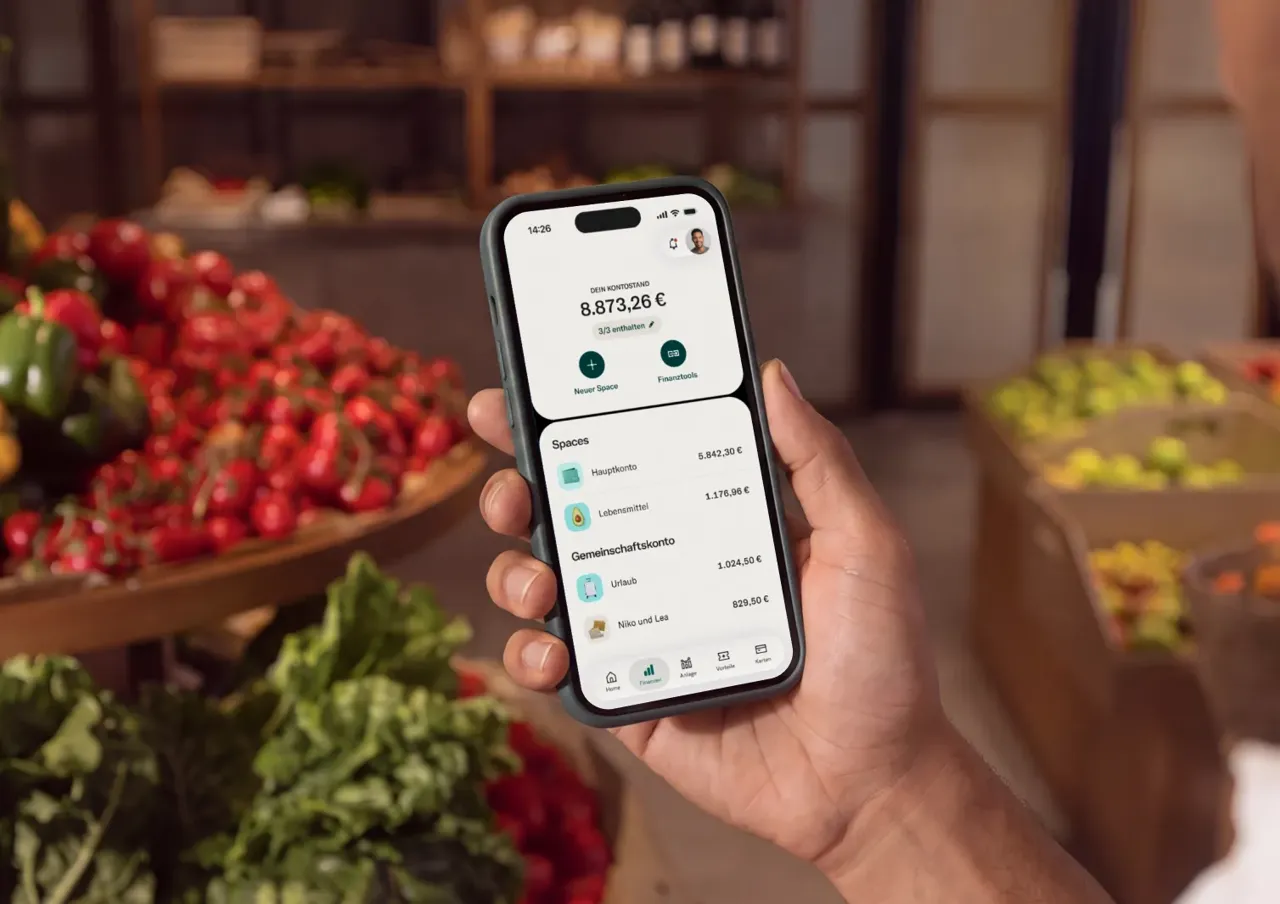 Eine Hand hält ein Smartphone mit der Benutzeroberfläche einer Banking-App, die Unterkonten und Kontostände vor einem verschwommenen Ladenhintergrund zeigt.