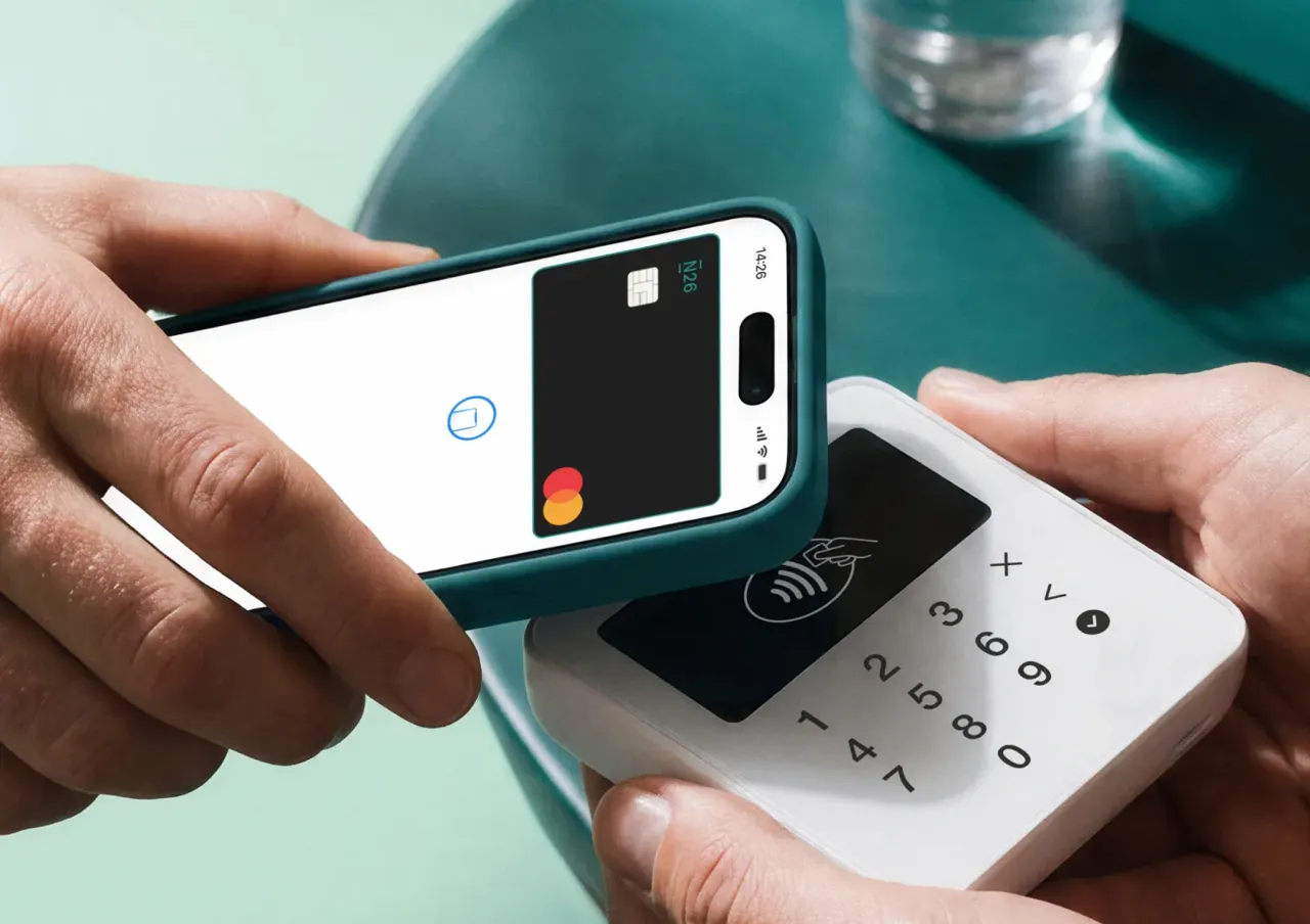A hand holds a smartphone displaying the N26 app card settings screen with an Add to Apple Wallet button, resting beside a glass of water on a teal table.