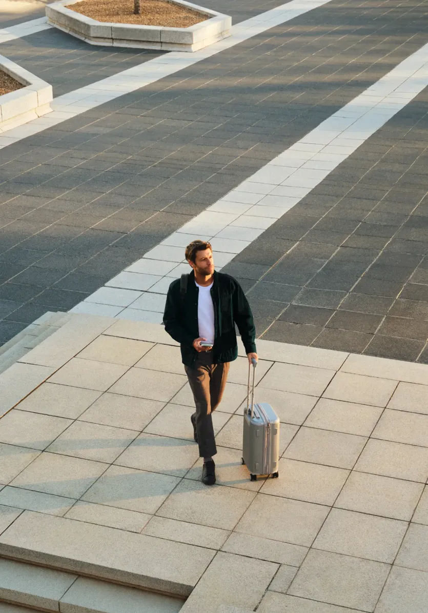 A man walks across a paved outdoor plaza pulling a rolling suitcase behind him.