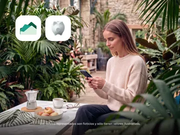 Una mujer utiliza su teléfono inteligente sentada en la terraza de un jardín junto a una cafetera y fruta, con iconos de la aplicación de inversión flotando arriba.
