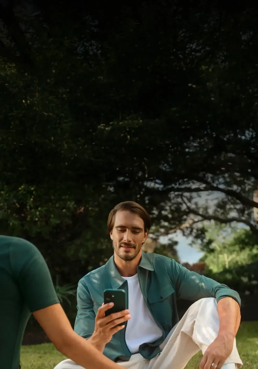 A man sits on a blanket outdoors in a park and looks at his smartphone screen beside another person.