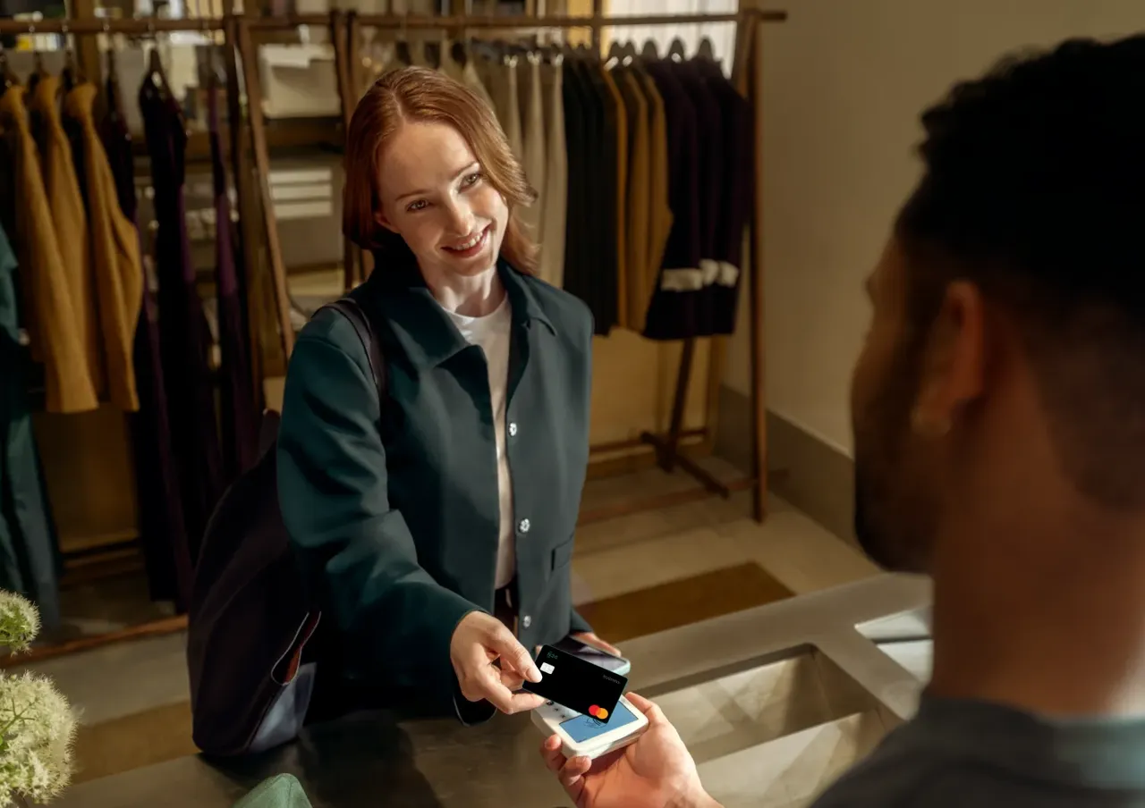 A smiling woman pays for a purchase by tapping a dark credit card on a contactless terminal held by a store associate in a clothing boutique.