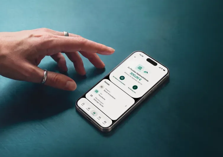Eine Hand liegt auf einer dunkelblau-grünen Oberfläche neben einem Smartphone, das die Details einer Transaktion von 300,00 € auf ein Geldmarktkonto anzeigt.