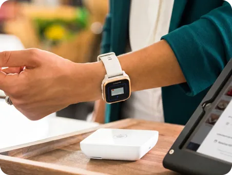 A close-up photograph shows a person’s arm with a smartwatch displaying a card held above a contactless payment terminal.