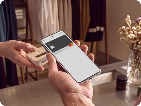 A first-person view photograph from behind a person's shoulder as they hold a smartphone up to a wireless payment terminal being presented by another person's hand.