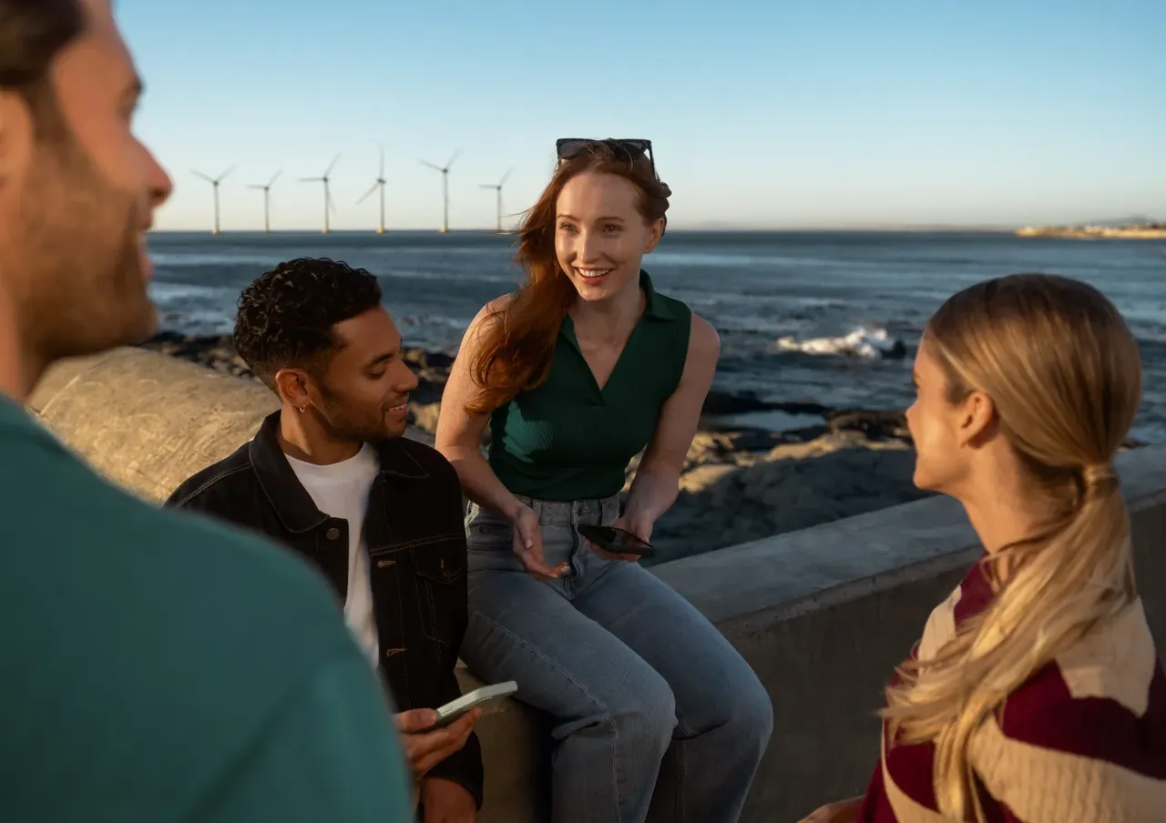 Drei Personen unterhalten sich im Freien an einer Küstenmauer mit Windrädern auf dem Meer im Hintergrund.