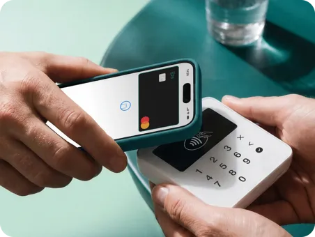 A close-up photograph shows one person's hands holding a green-cased smartphone to a wireless payment terminal held by another person's hands.