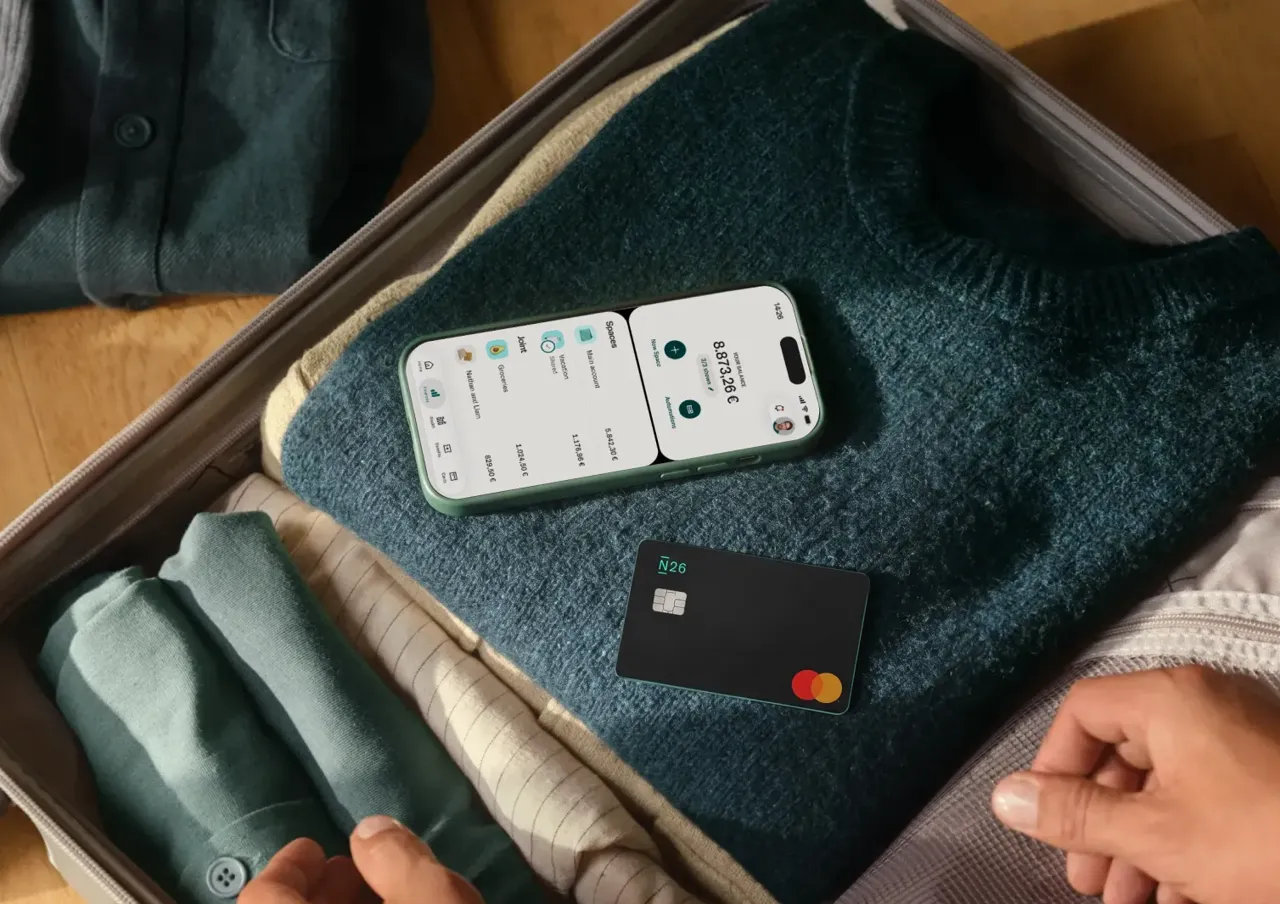 A smartphone displaying a banking app and a black N26 payment card rest on a folded sweater inside an open suitcase.