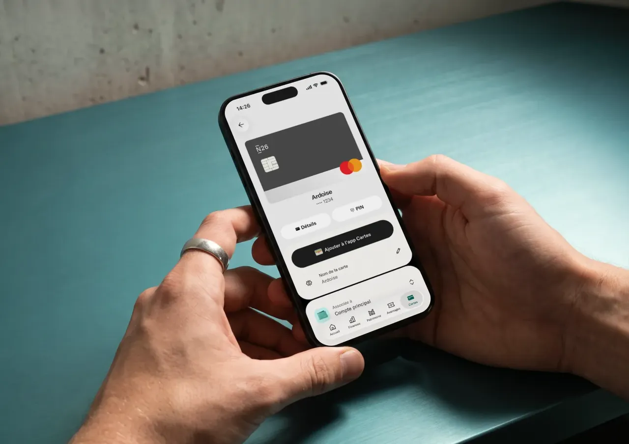 Deux mains tiennent un smartphone affichant un écran de gestion de carte N26 avec l'option Ajouter à Apple Wallet.