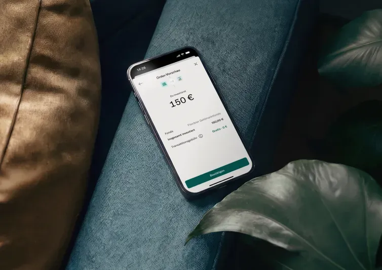 Ein Smartphone mit der Bestellvorschau einer Finanz-App zur Einzahlung von 150 € auf ein Geldmarktkonto liegt auf einem dunkelblauen Kissen neben einem Pflanzenblatt.