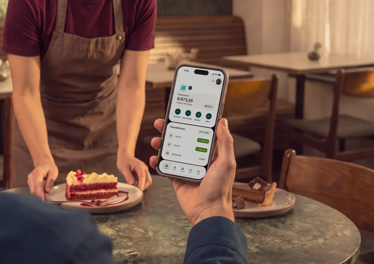 Eine Person hält ein Smartphone mit der Benutzeroberfläche einer Banking-App mit einem Kontostand von 8.873,26 € in der Hand, während sie an einem Cafétisch gegenüber einer Bedienung sitzt, die ein Dessert serviert.
