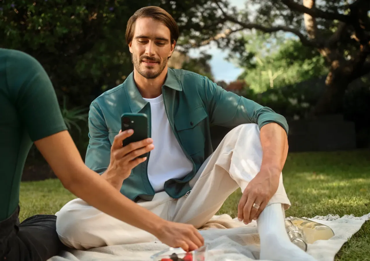 Un hombre está sentado sobre una manta al aire libre y mira un smartphone que tiene en la mano, junto a otra persona y unos aperitivos.