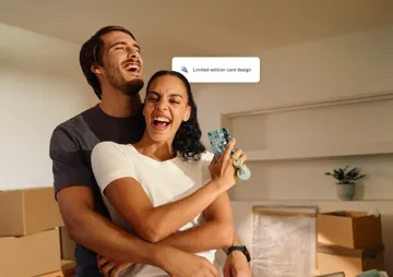A laughing young couple embraces in a room filled with moving boxes, while the woman holds up a colorful credit card and a set of keys.