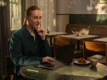 A woman sitting at a café table works on a laptop next to a cup of coffee and a pastry.