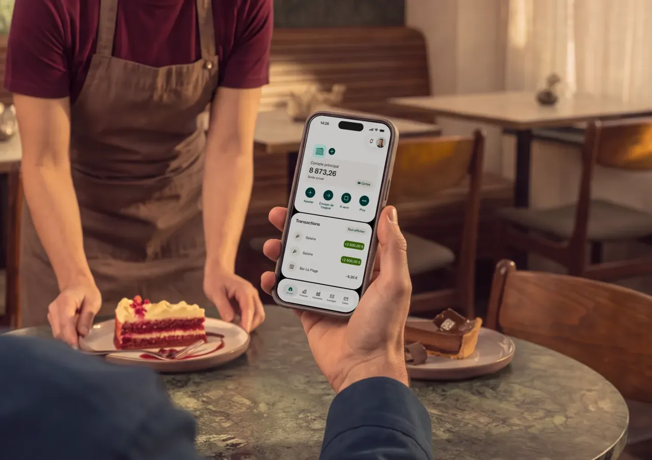 Une personne tient un smartphone affichant l'interface d'une application bancaire avec un solde de 8 873,26 € tout en étant assise à la table d'un café face à un serveur apportant un dessert.