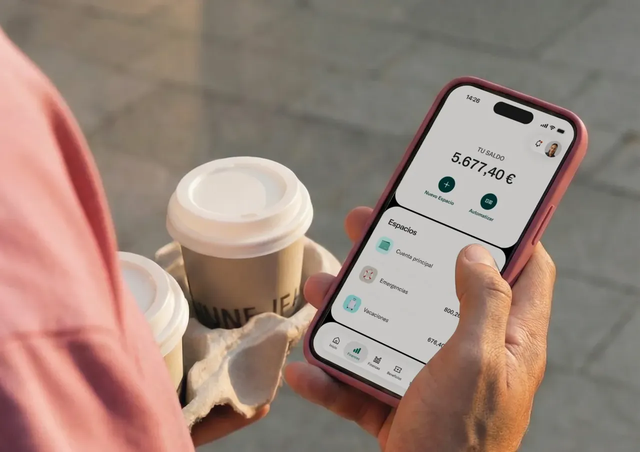 Una persona que lleva una bandeja de cartón con dos tazas de café para llevar utiliza una aplicación bancaria en un teléfono inteligente.