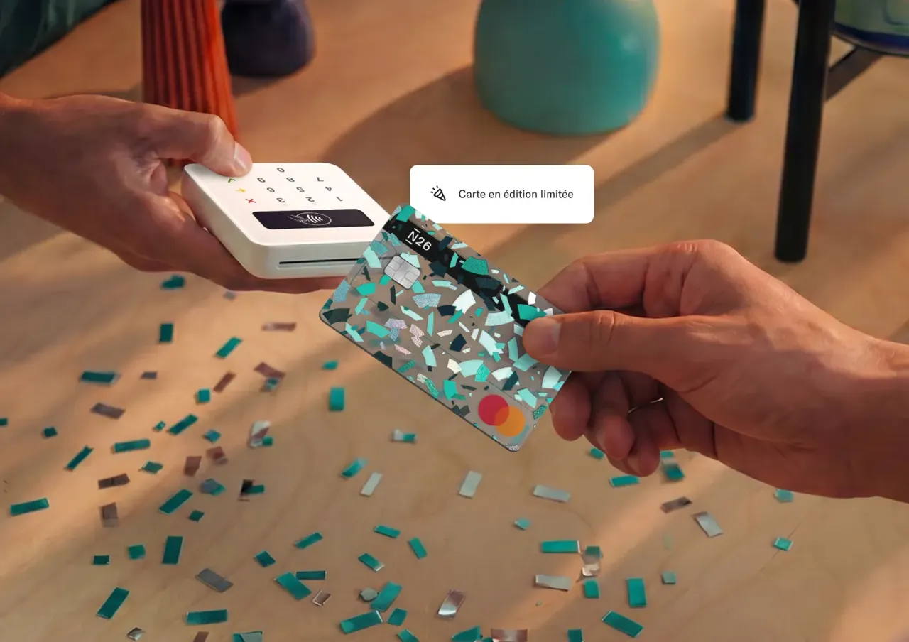 Vue en gros plan d'une personne utilisant une carte Mastercard N26 en édition limitée à motif confettis pour effectuer un paiement sans contact sur un terminal blanc au-dessus d'une table parsemée de confettis.