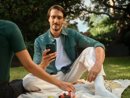 A man sits on a blanket during an outdoor picnic looking at his smartphone.