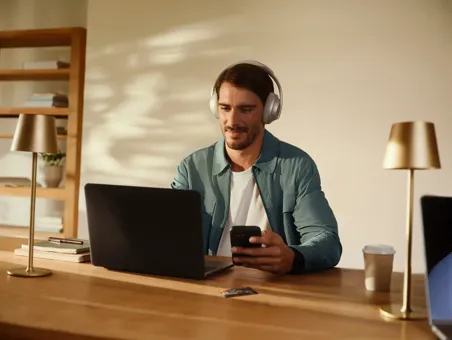 A man wearing headphones works at a wooden desk with a laptop while holding a smartphone.